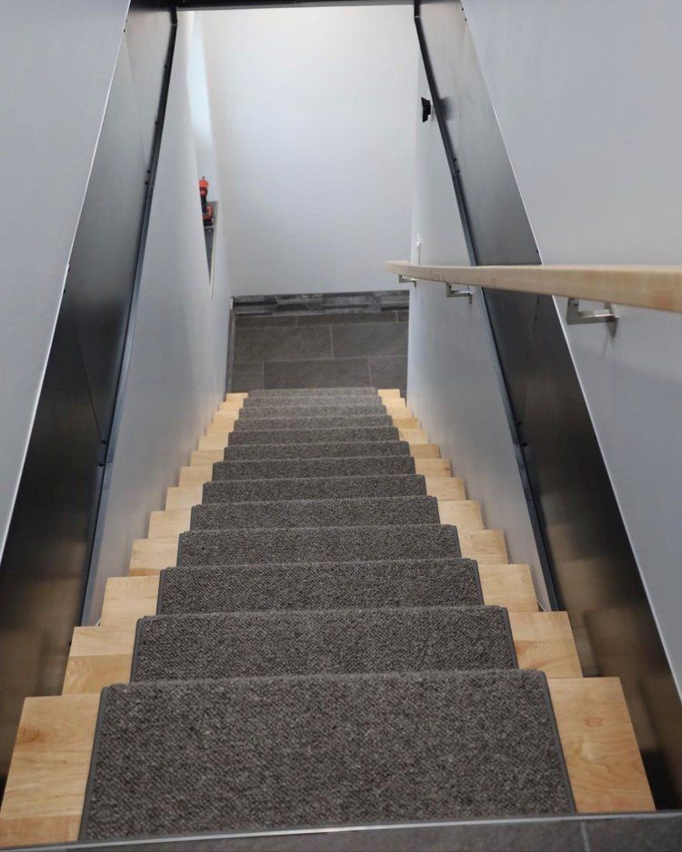 Staircase descending downwards. Gray carpet runner on wooden steps. Gray walls with metal trim. Handrail on the right.