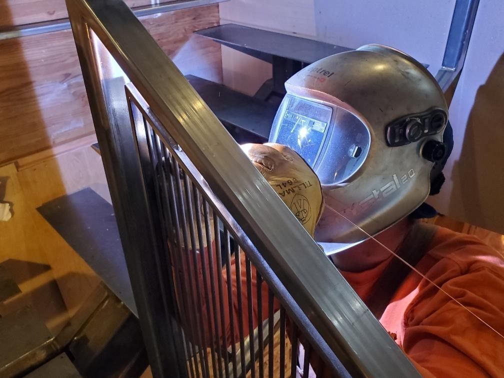 Welding the Outdoor Railings — Seattle, WA — BSMWI Welder wearing a helmet working on metal railing in a home, orange shirt visible.