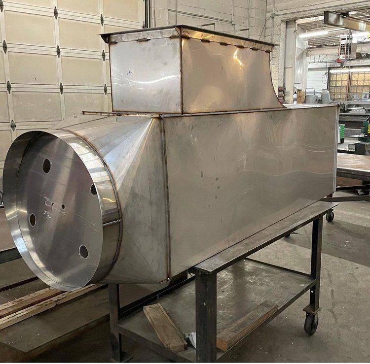 Stainless steel industrial ductwork resting on a metal cart in a workshop setting.
