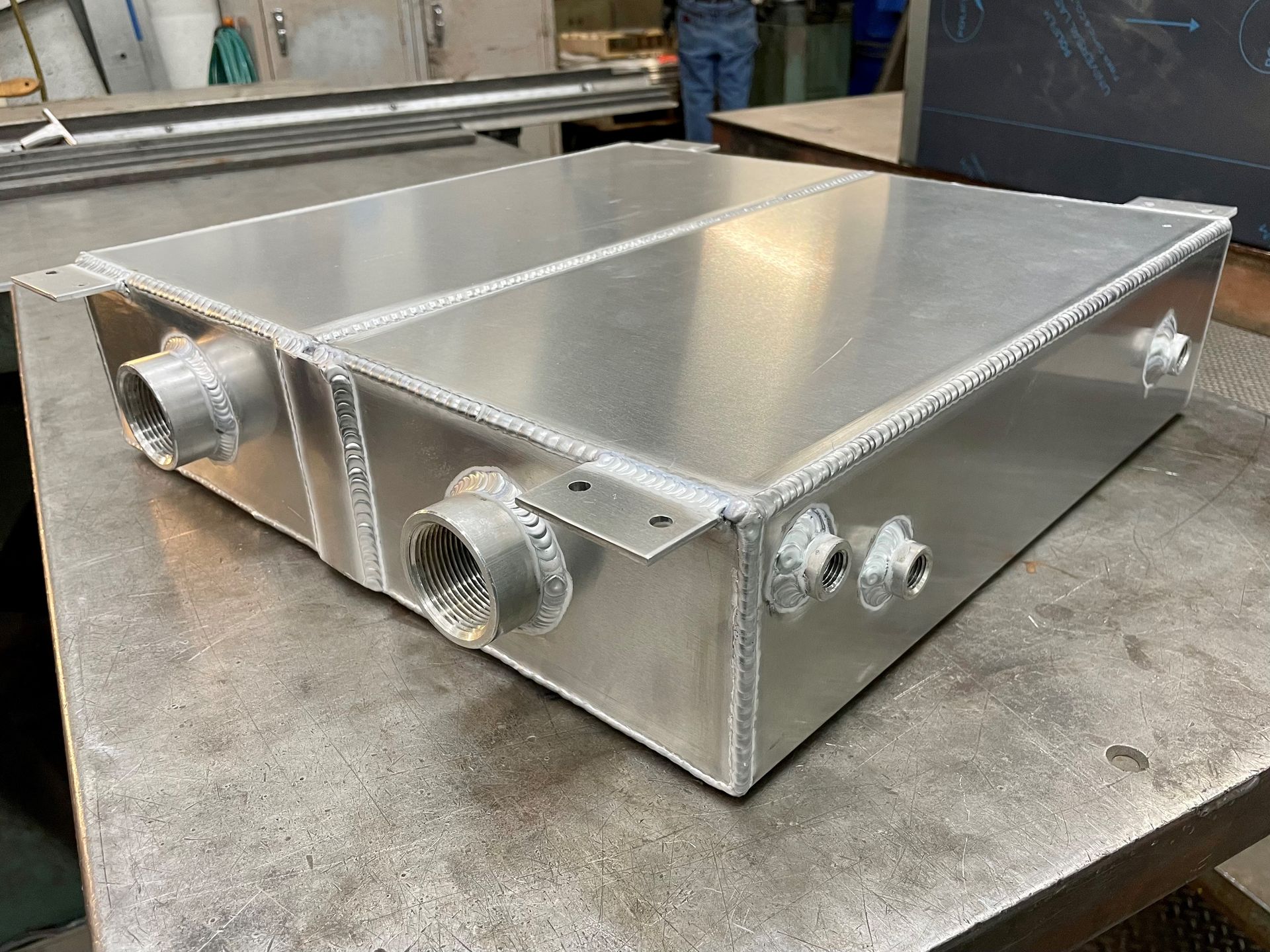 A rectangular, aluminum tank with welded seams and pipe fittings sits on a metal surface.