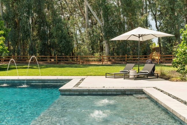 Backyard pool with lounge chairs, umbrella, and trees behind a wooden fence