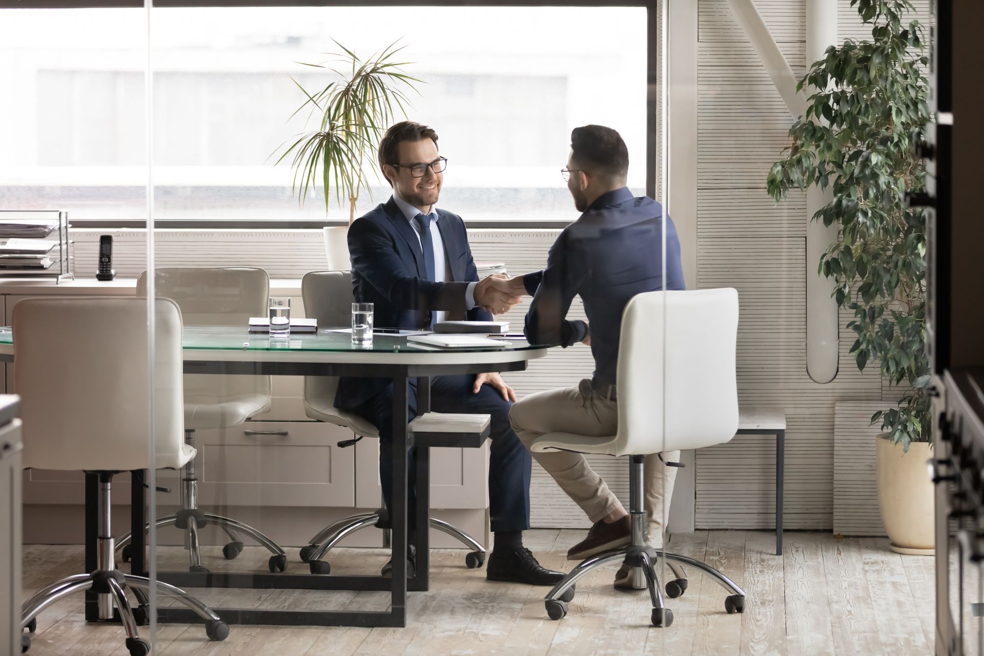 Two men in suits meet in a bright office, seated across a table near a large window.