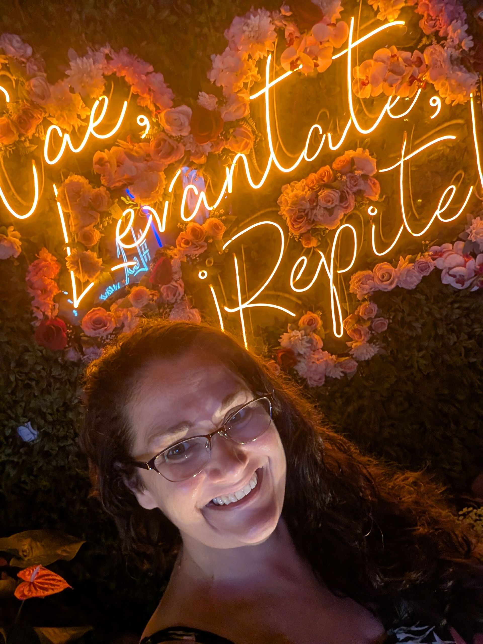 Woman smiling in front of a neon sign with Spanish text; sign is surrounded by flowers.