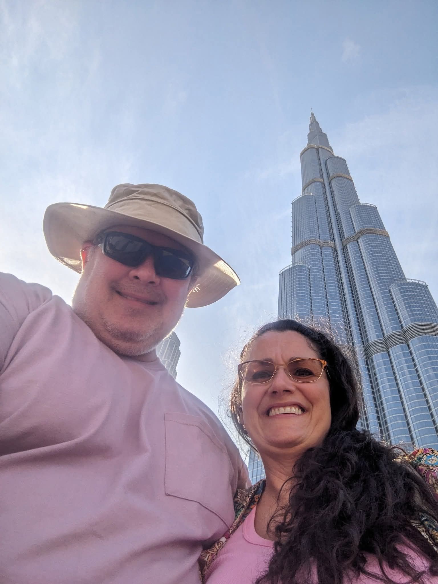 Couple smiles in front of the Burj Khalifa, Dubai. The man wears a hat and sunglasses, woman wears glasses.