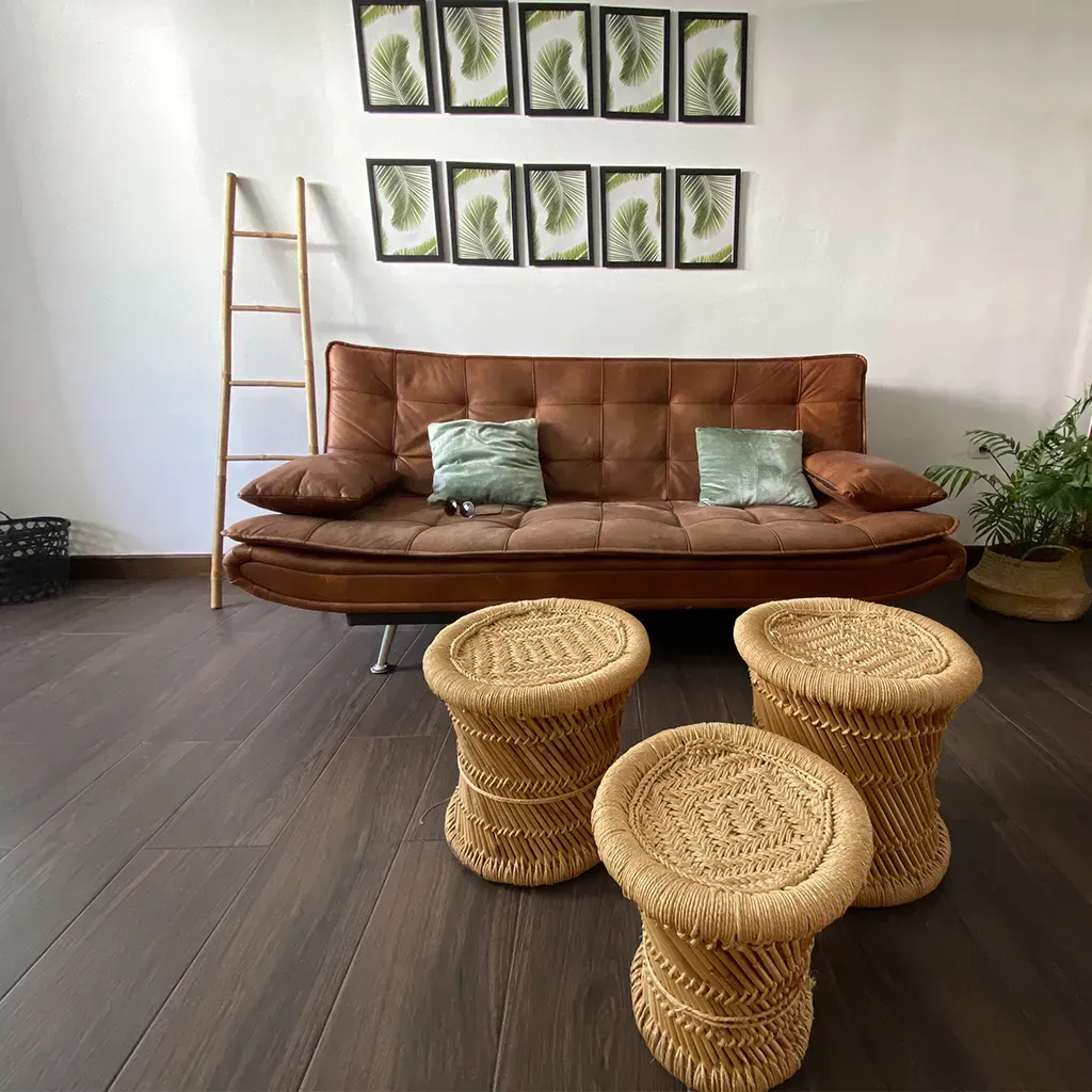 Acogedor rincón de salón con sofá marrón, taburete de mimbre, escalera y cesta sobre suelo de madera gris.
