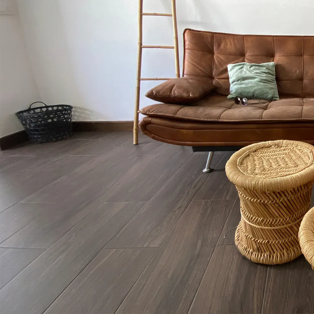 Acogedor rincón de salón con sofá marrón, taburete de mimbre, escalera y cesta sobre suelo de madera gris.