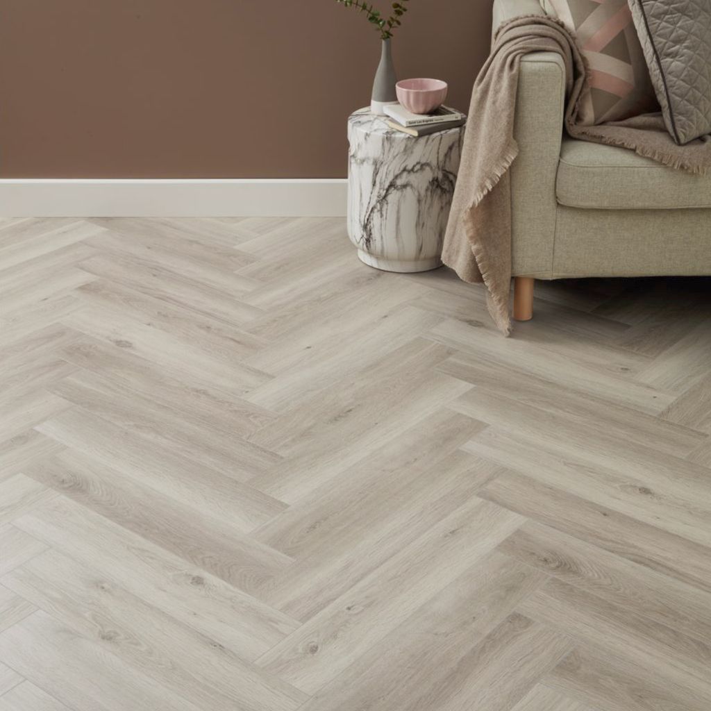 Suelo de madera clara en espiga junto a un sofá beige, con una mesa auxiliar de mármol y decoración rosa.