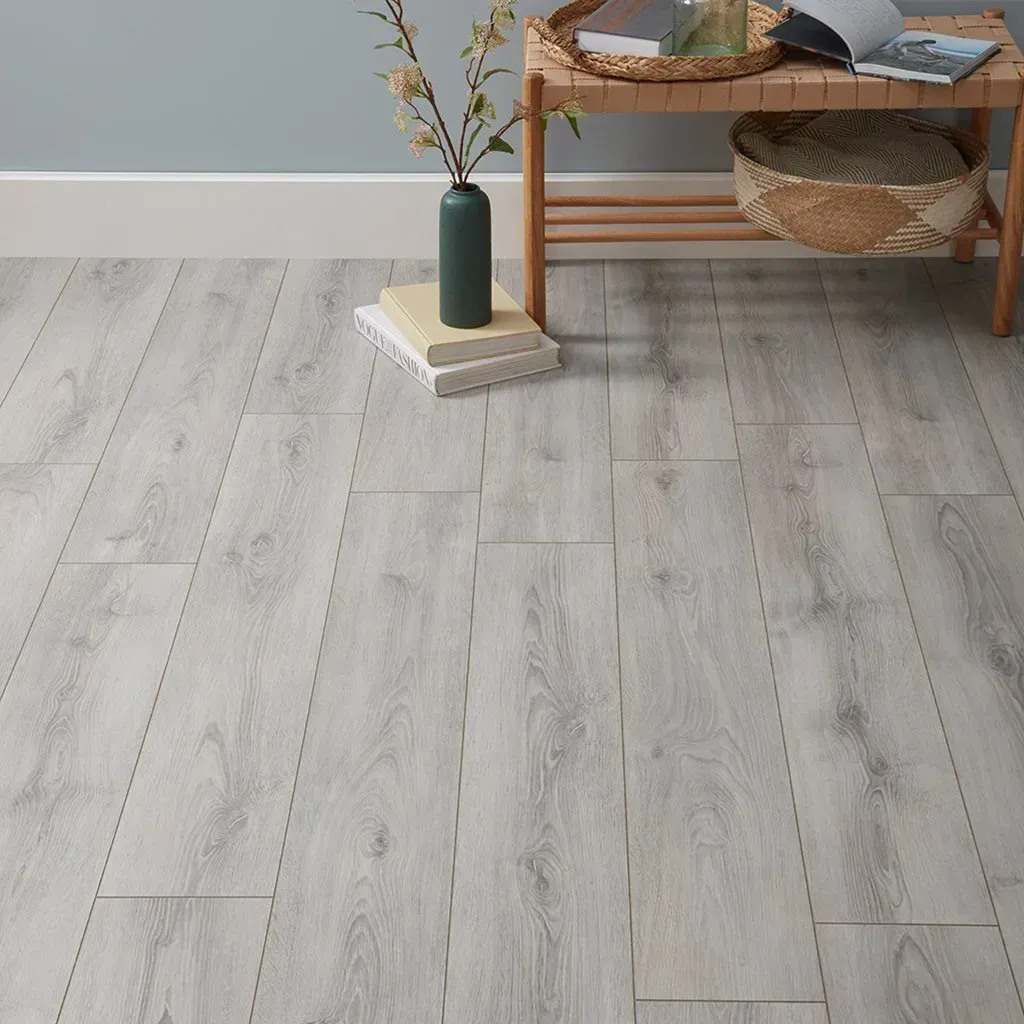 Suelo de madera gris claro con una mesita que sostiene un jarrón y una cesta, junto a una silla y una pila de libros.