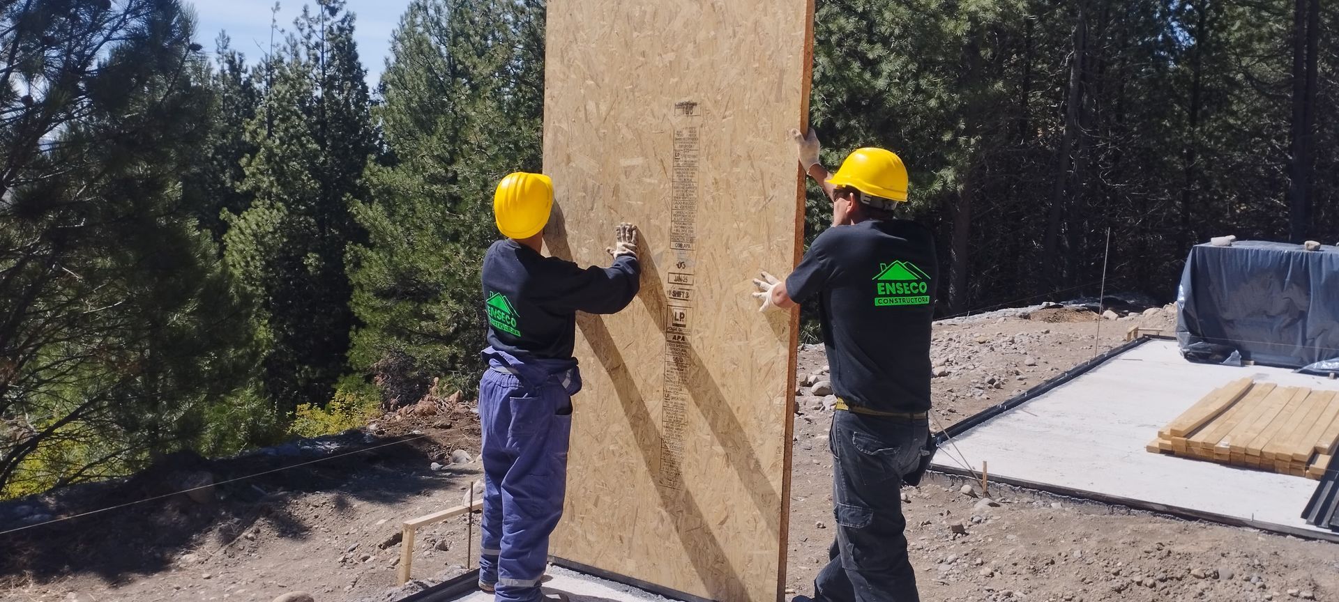 Dos trabajadores con cascos amarillos aseguran un panel de madera al aire libre en una obra de construcción.