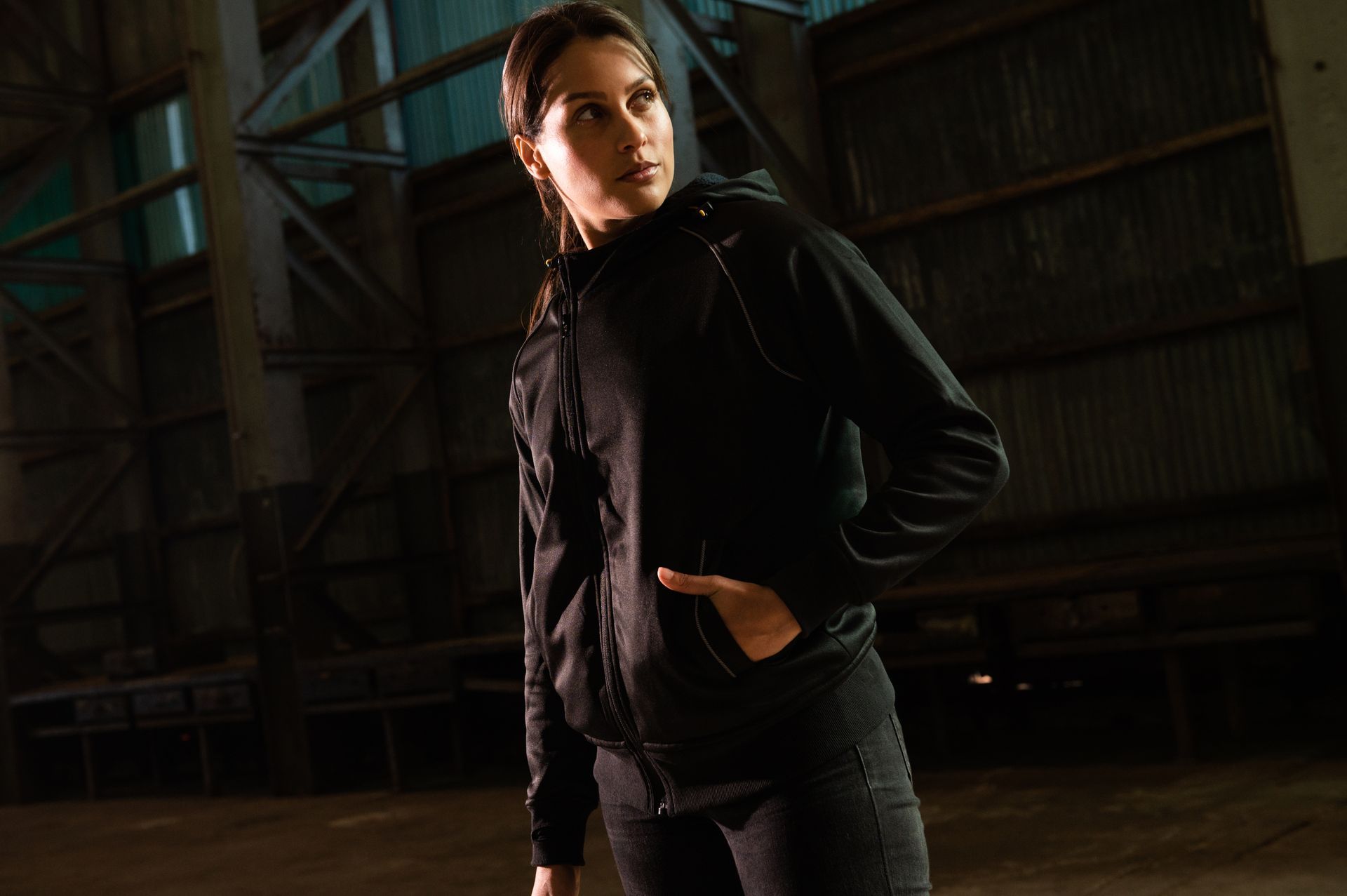 A woman in a black jacket is standing in a dark room with her hands in her pockets.