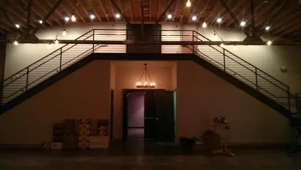Interior view of a building with a staircase leading to a mezzanine. Overhead lights, dark door.