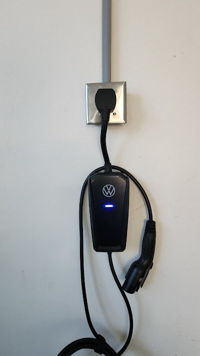 Black EV charger plugged into a wall outlet on a white wall, emitting blue light.