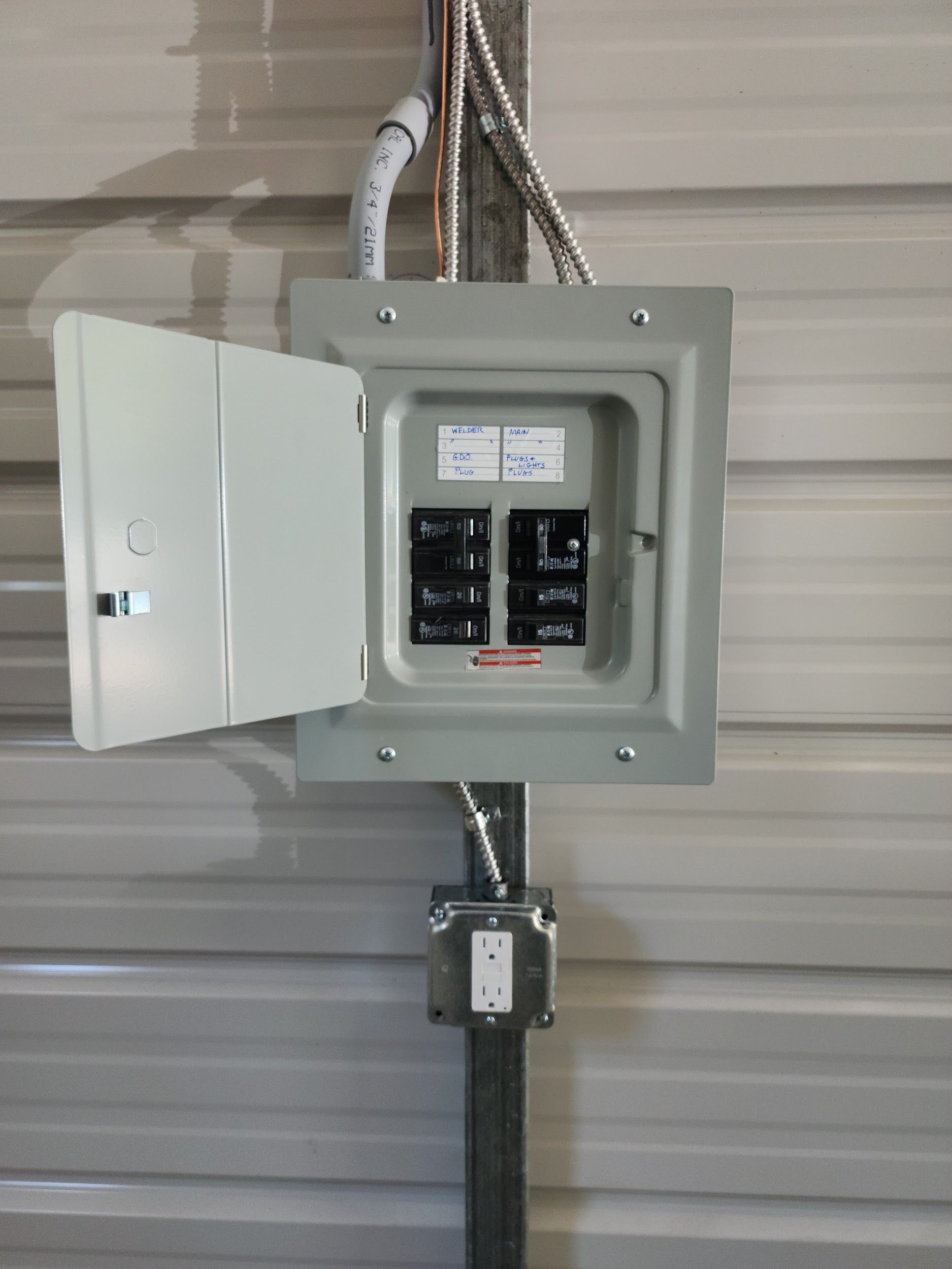 Electrical panel with the door open, mounted on a metal wall. Below is a grounded outlet.