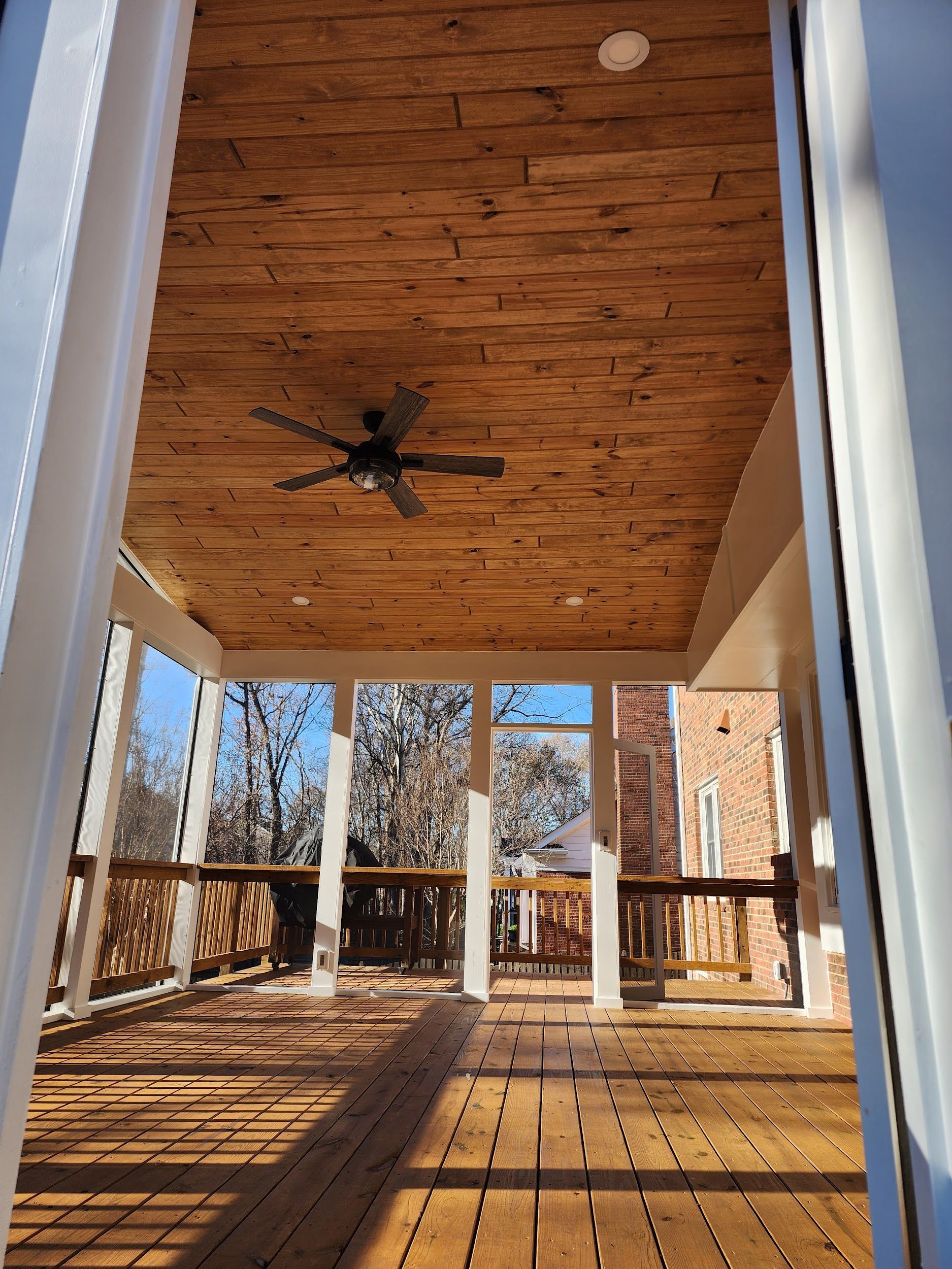 Covered wooden deck with a wood ceiling, ceiling fan, and white support columns.