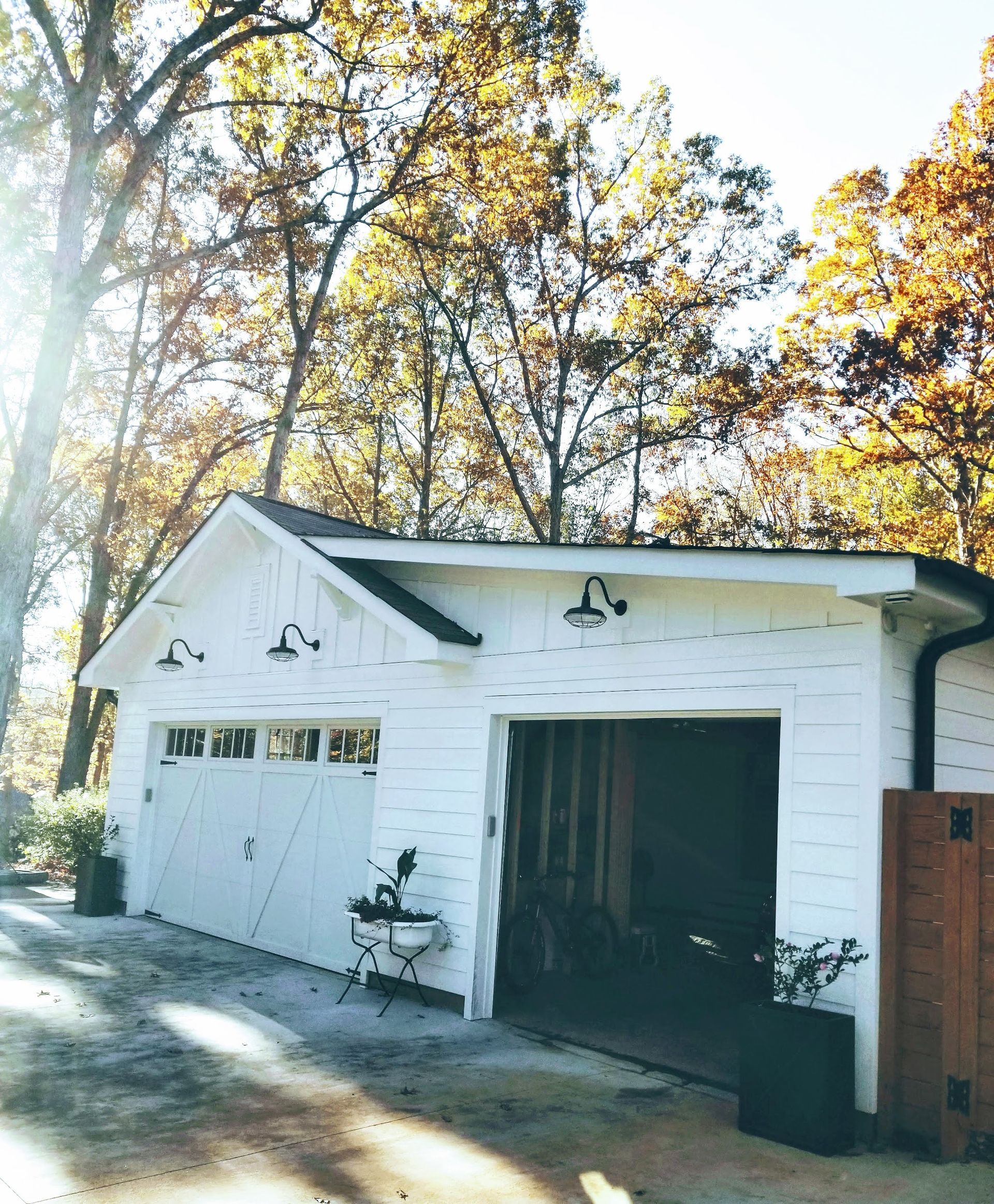 White two-car garage with open door, black trim, and barn lights, set against autumnal trees.