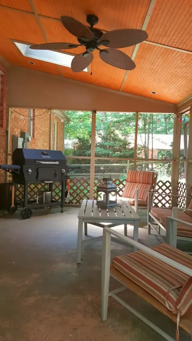 Screened porch with grill, seating, and ceiling fan. Orange ceiling, brick wall, and trees visible.