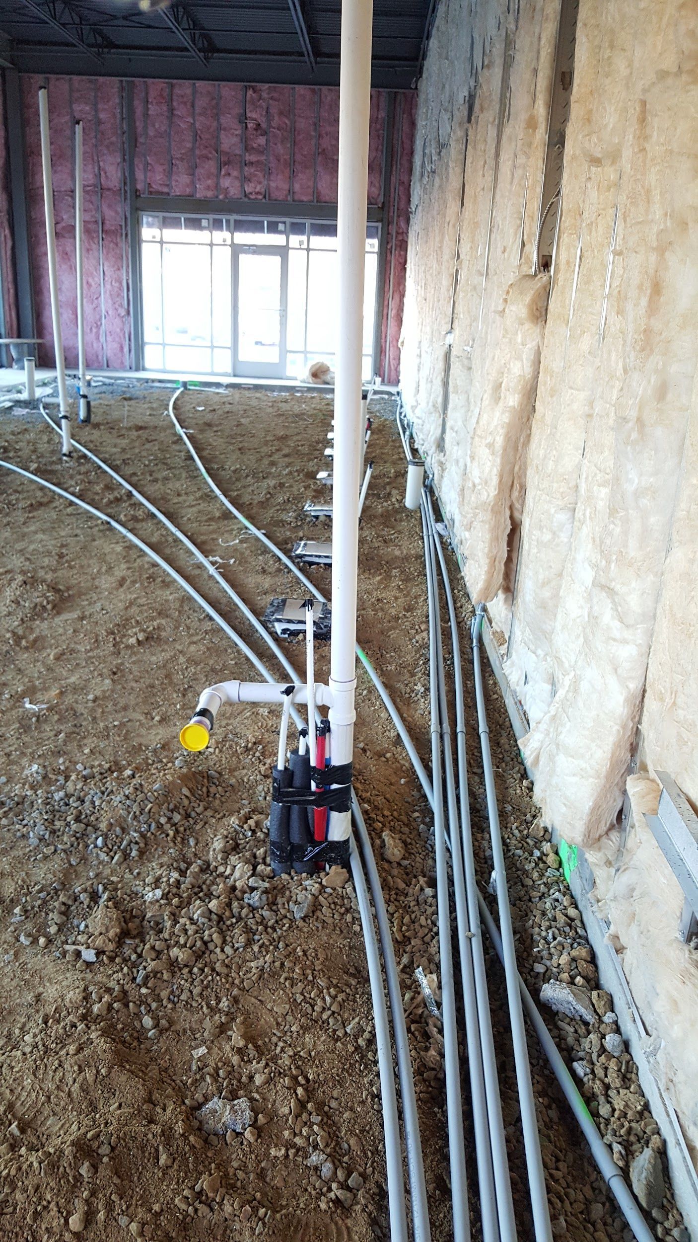 Construction site with exposed plumbing and wiring; pipes run along a dirt floor next to an insulated wall.