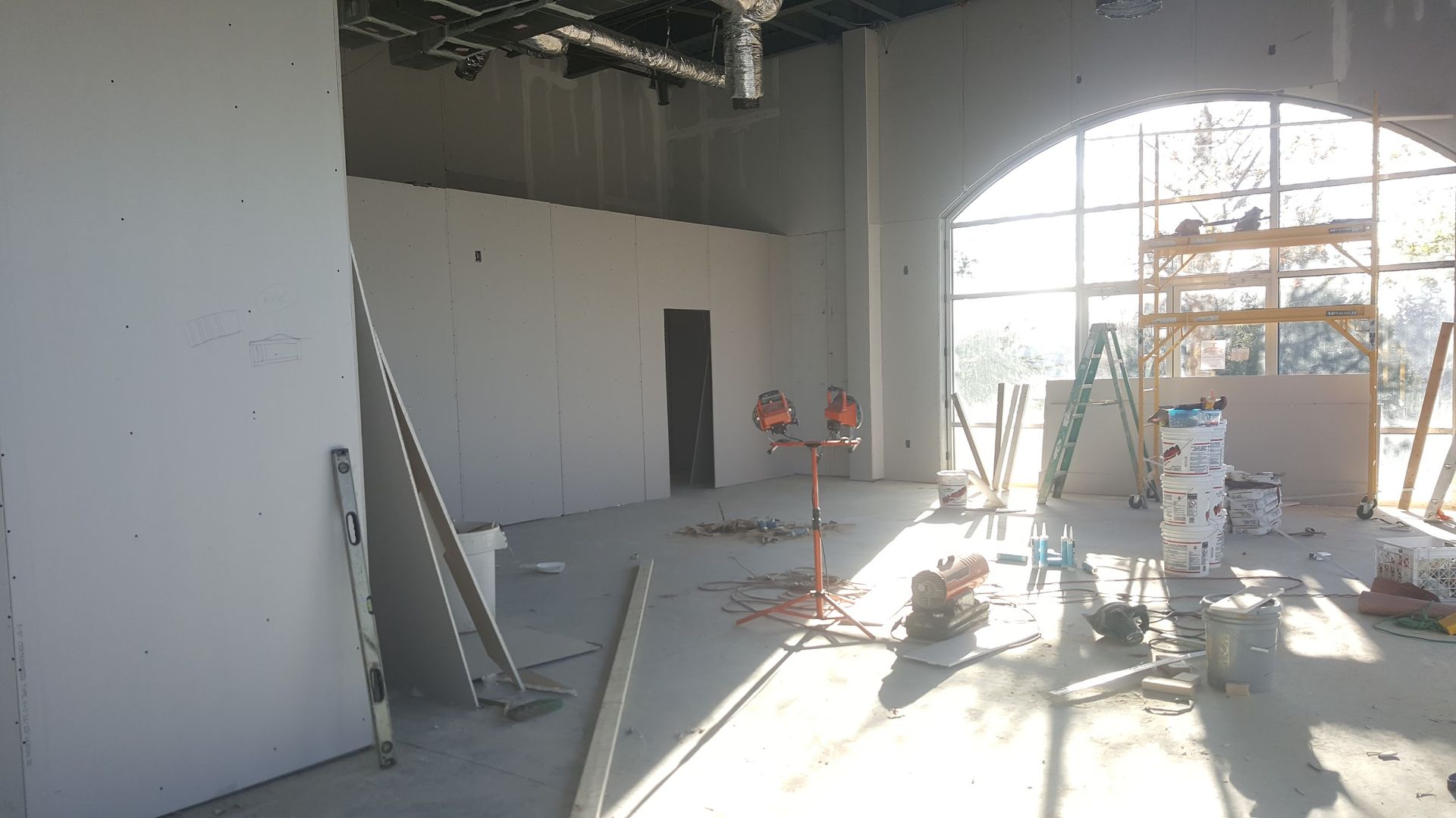 Interior construction site with drywall on walls and tools scattered about. Large arched window lets in sunlight.
