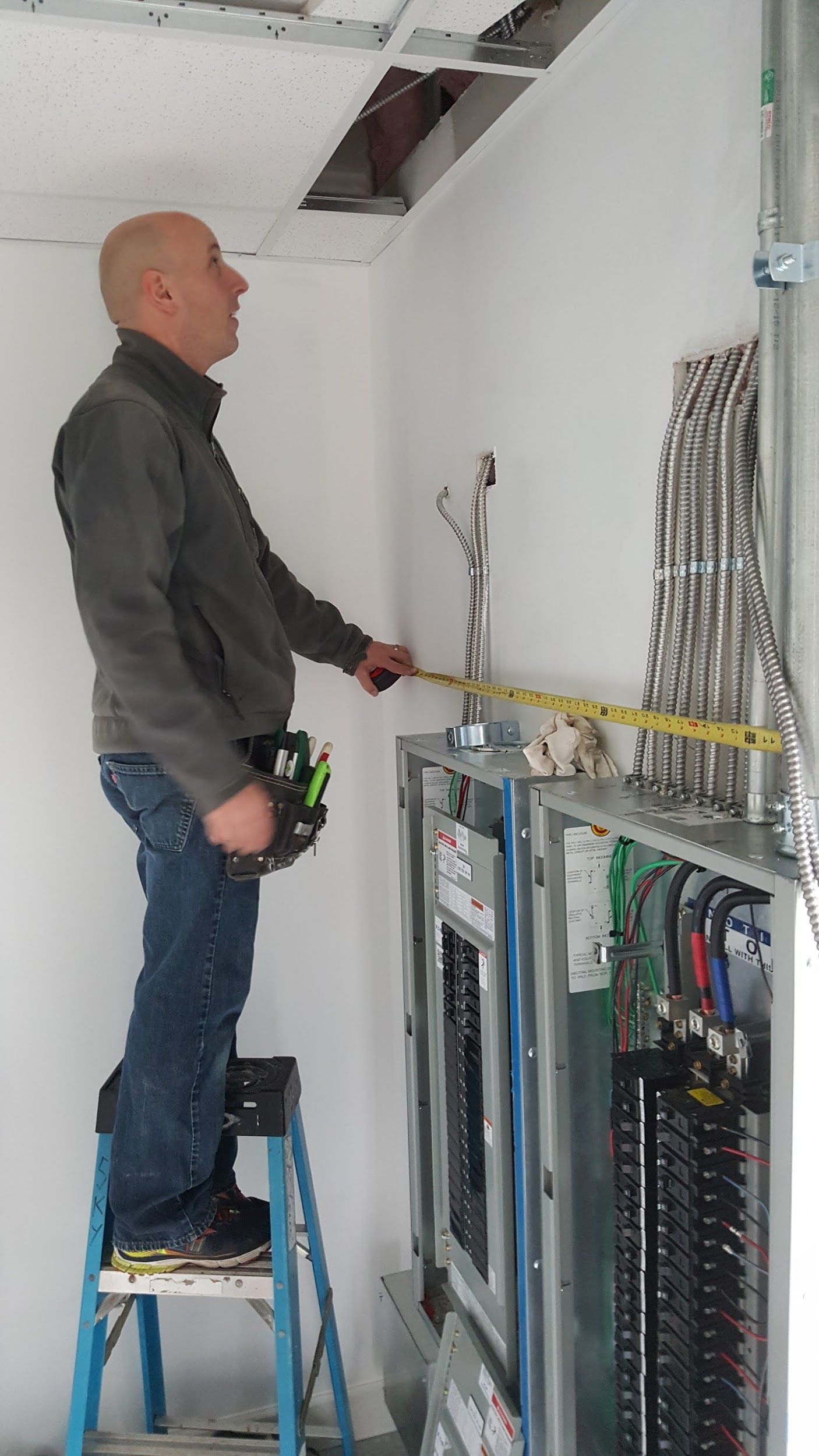 Electrician on a stepladder measures a circuit breaker panel in a building, using a tape measure.