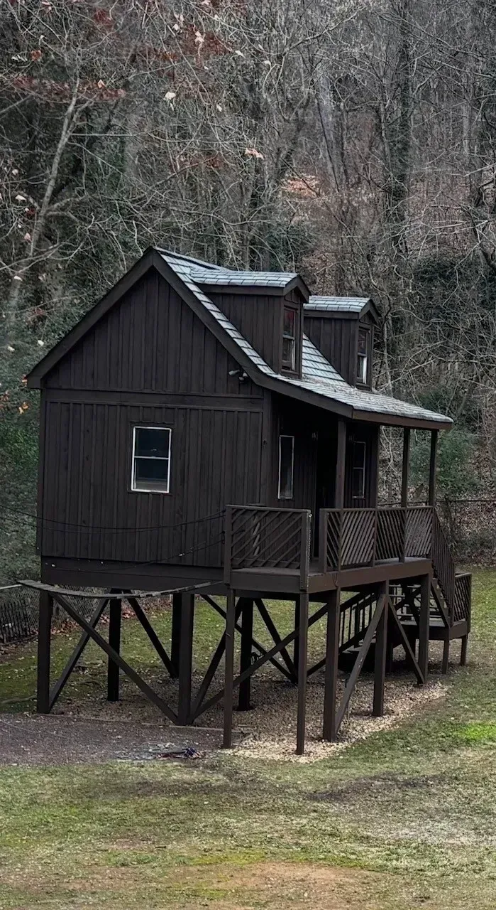 Small black wooden cabin on stilts in a wooded area, with a porch and stairs.