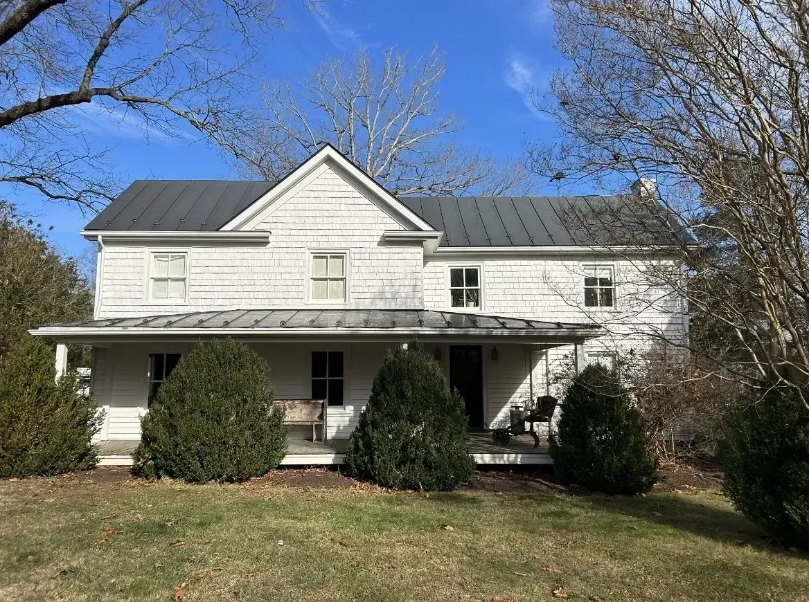 White two-story house with a front porch, dark roof, and shrubs under a blue sky