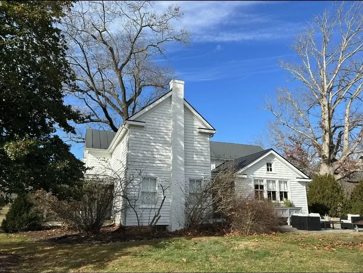 White house with chimney and black roof amid leafless trees under a blue sky