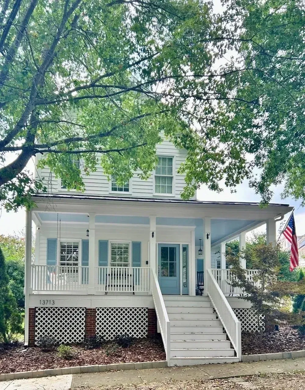 White house with front porch and steps, framed by large trees and an American flag.