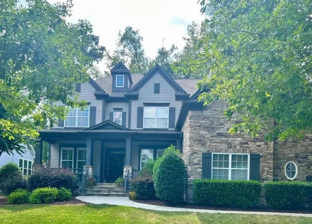 Stone house with dormers and a front porch, framed by trees and shrubs on a sunny day