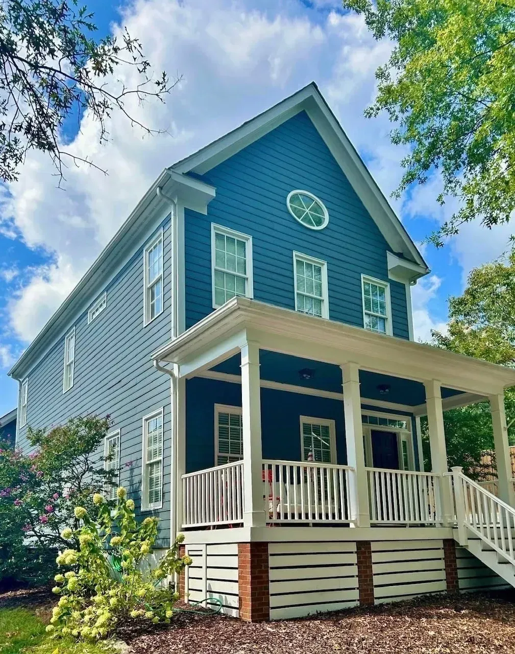 Blue two-story house with white porch and trim, viewed from below beneath leafy trees