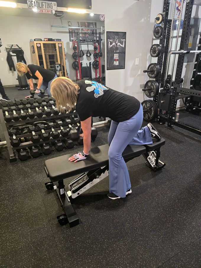Woman exercising, bent over on a bench in a gym, with one leg extended and hand supporting herself.