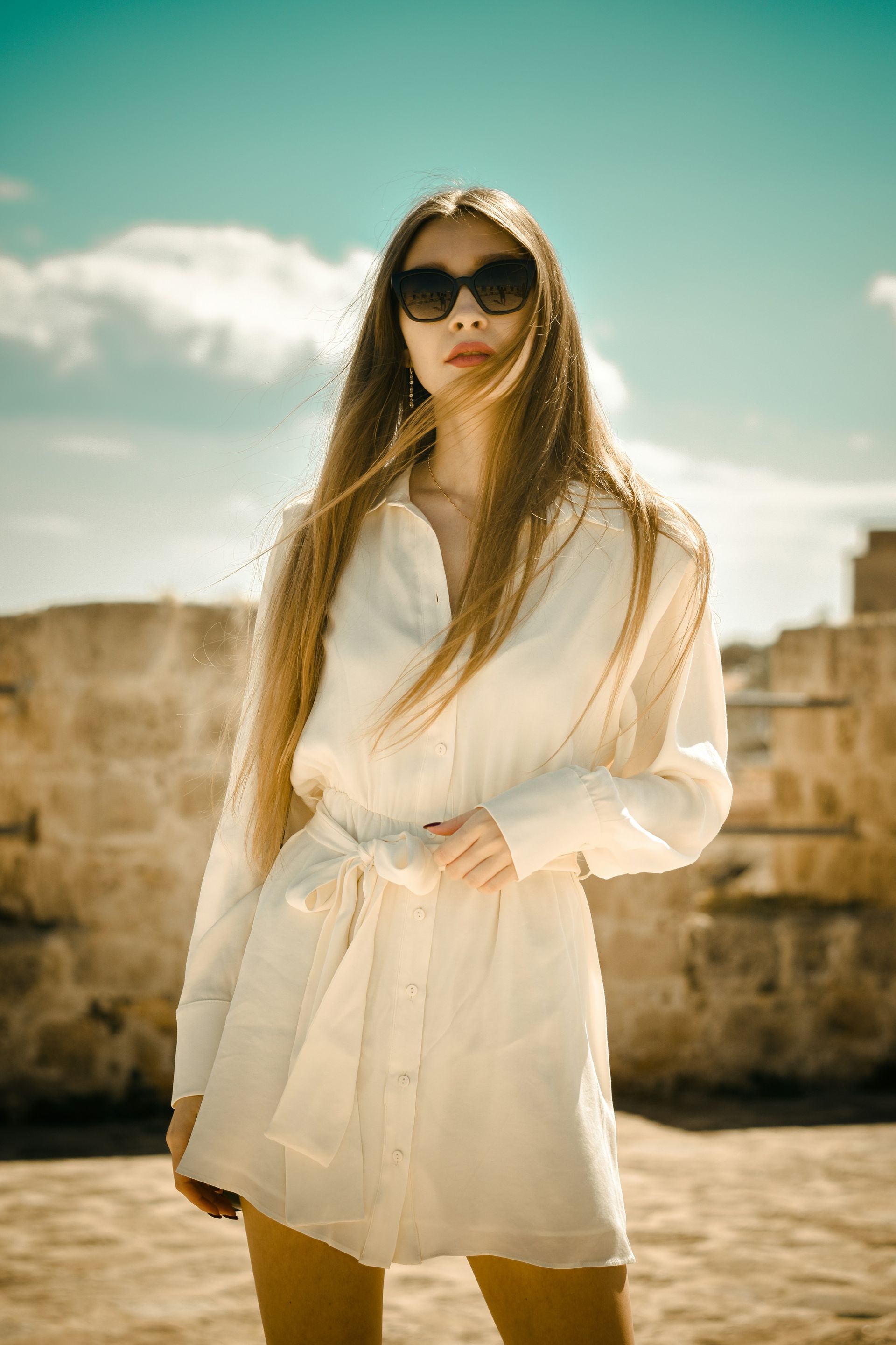 A person with long hair wearing sunglasses and a cream-colored, belted shirt dress, standing against a stone wall.