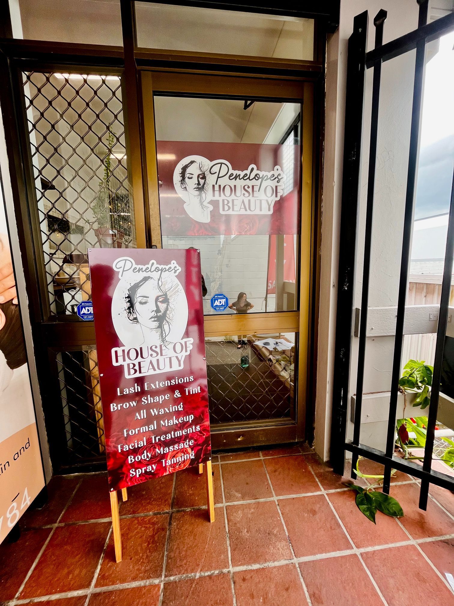 Entrance to a beauty salon. Maroon sign outside with services listed. Glass door with logo. Exterior, daytime.