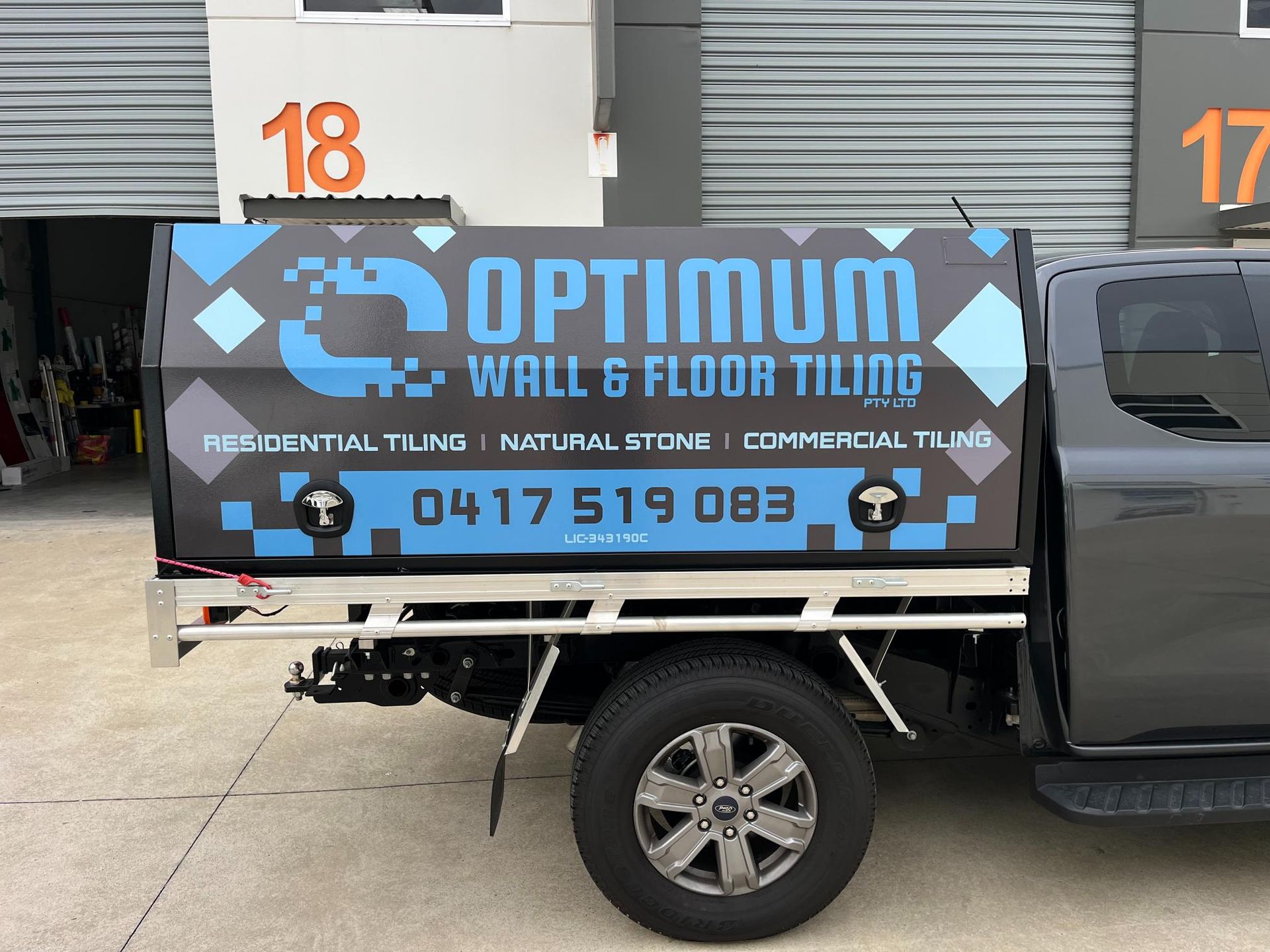 Gray truck with a customized bed promoting Optimum Wall & Floor Tiling services; contact number on display.
