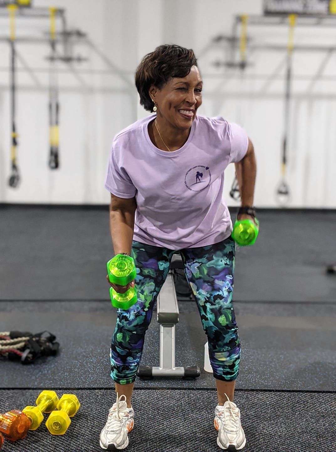 A woman is sitting on a bench in a gym holding green dumbbells.