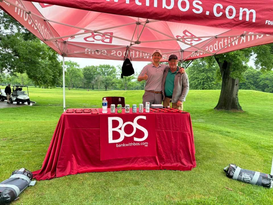 Two men are standing in front of a bos table on a golf course.
