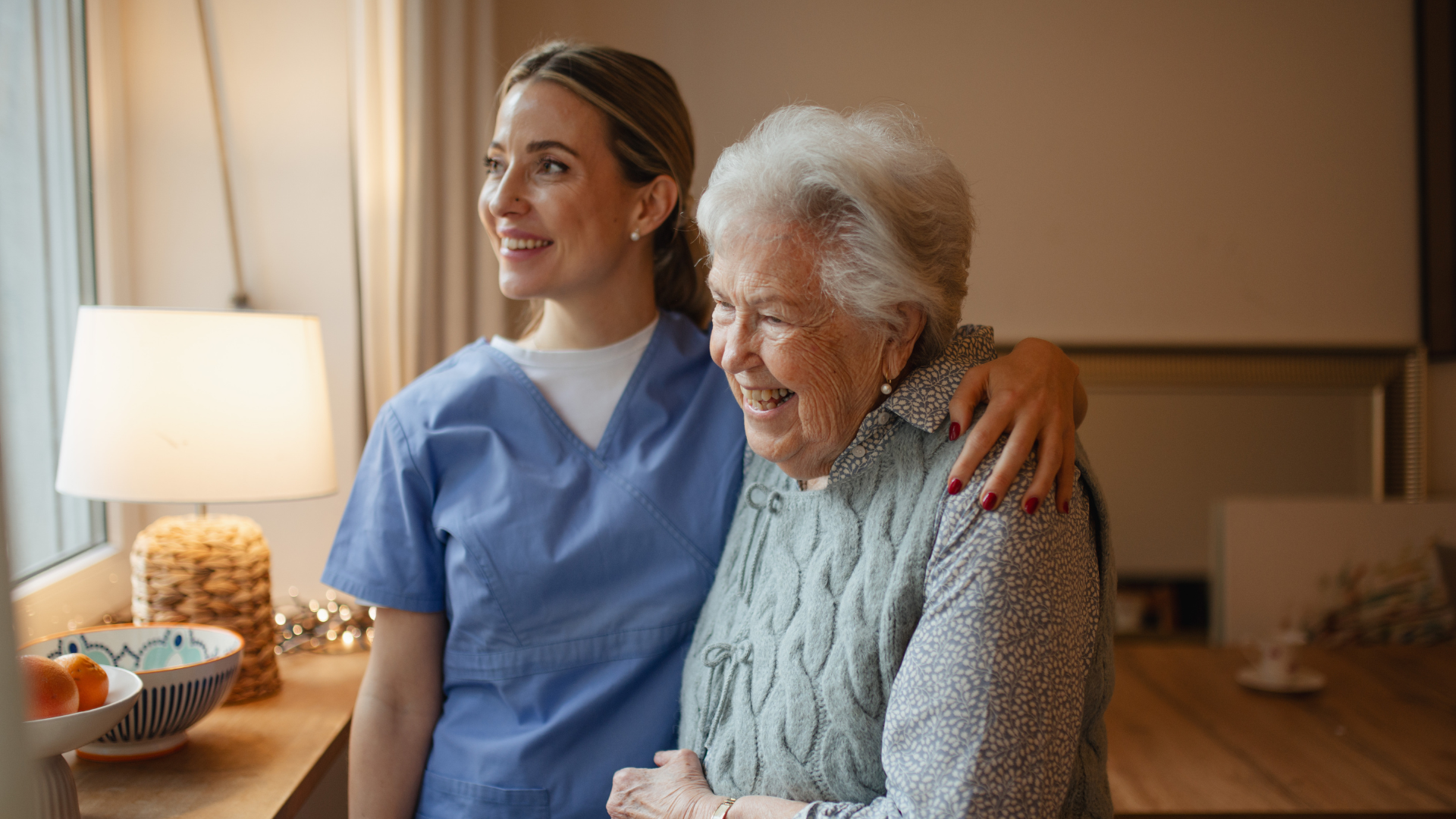 Nurse in blue scrubs smiling with an elderly woman in a care home room