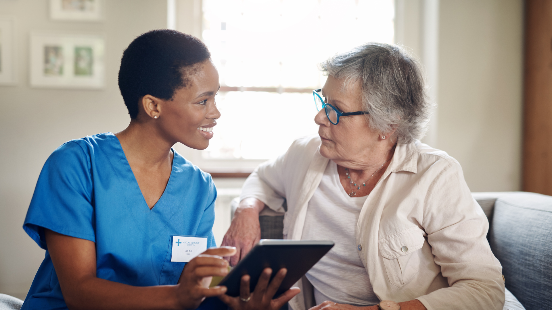 Caregiver talking with an older adult on a couch, holding a tablet in a bright living room.