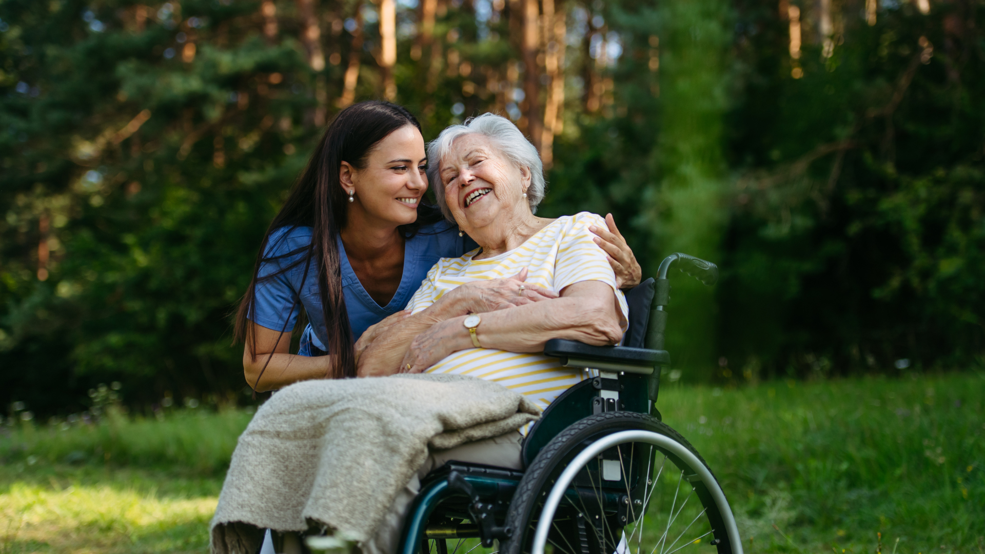 Woman leaning over and hugging an older person in a wheelchair outdoors on a grassy path.