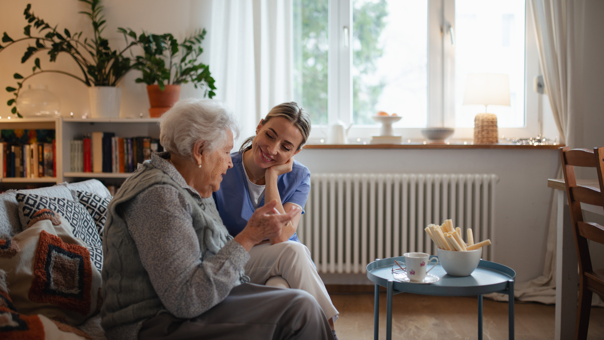 Caregiver talking with an elderly woman in a bright living room, with tea on a small table.