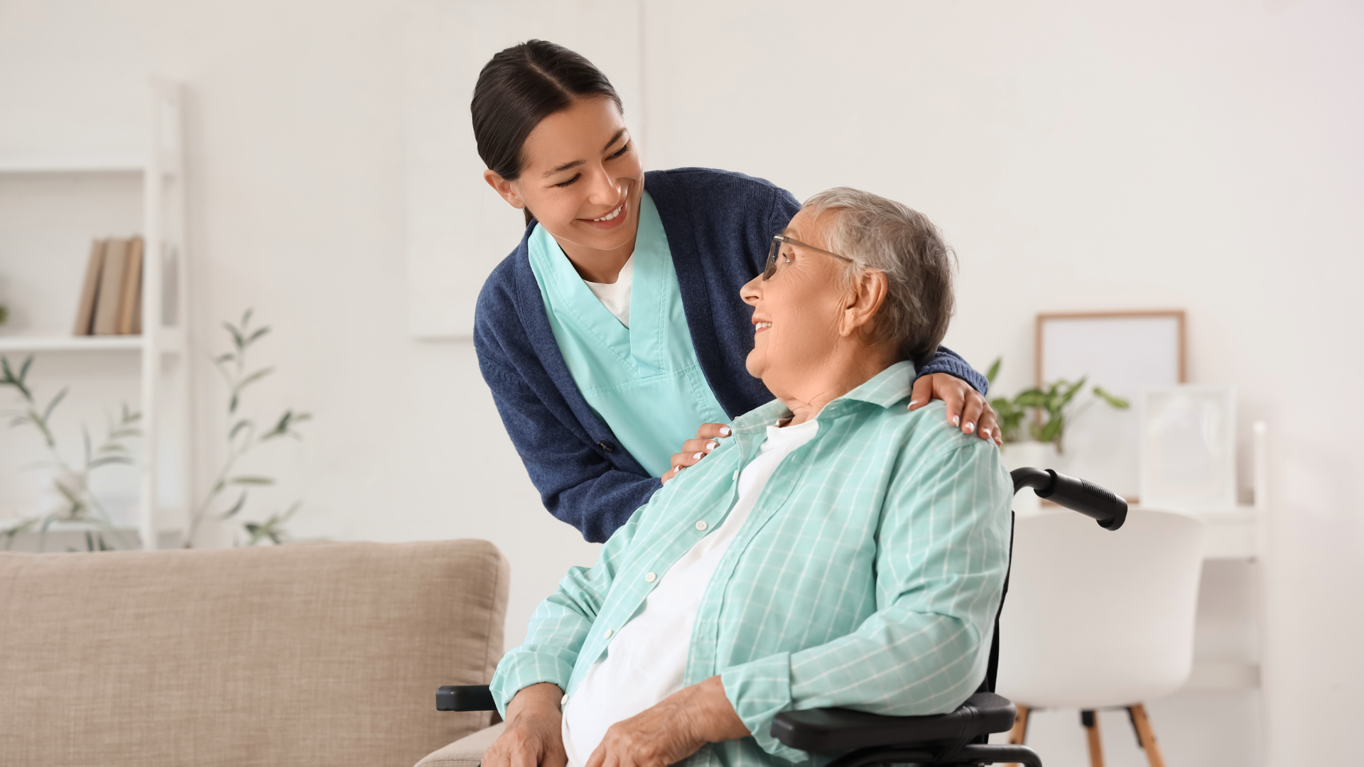 Caregiver leaning toward a seated older adult in a bright living room, sharing a warm conversation.