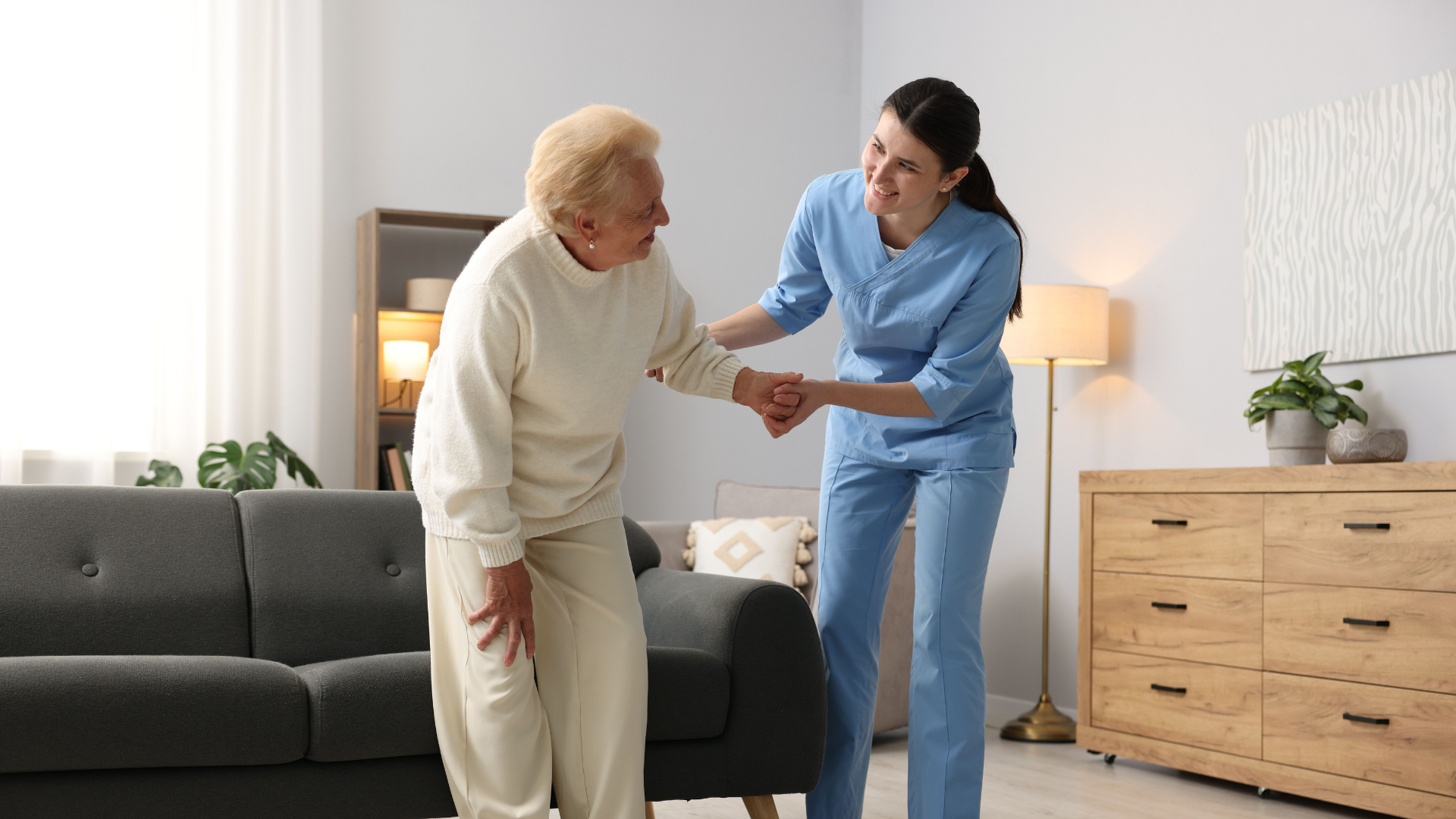 Caregiver helping a smiling elderly woman stand in a bright living room beside a gray sofa and wooden dresser