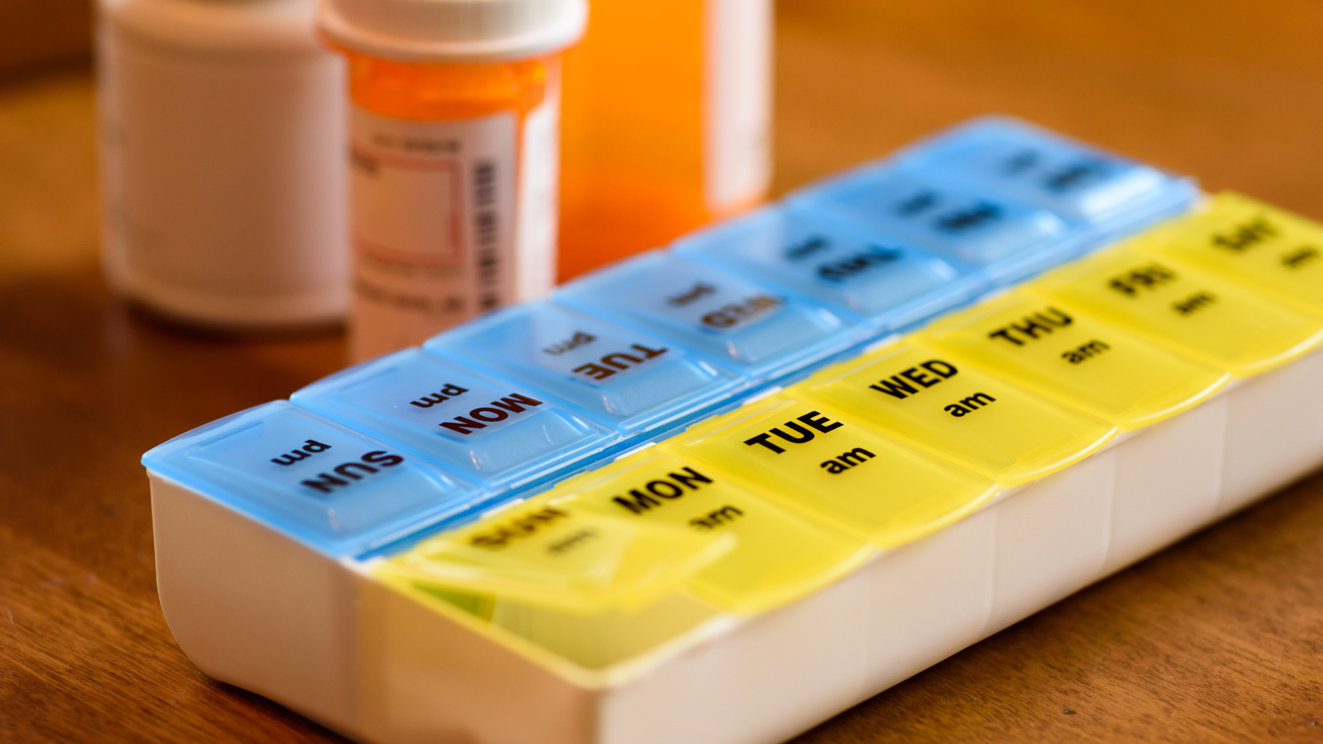 A blue and yellow pill organizer with labeled compartments beside a prescription bottle on a wooden table