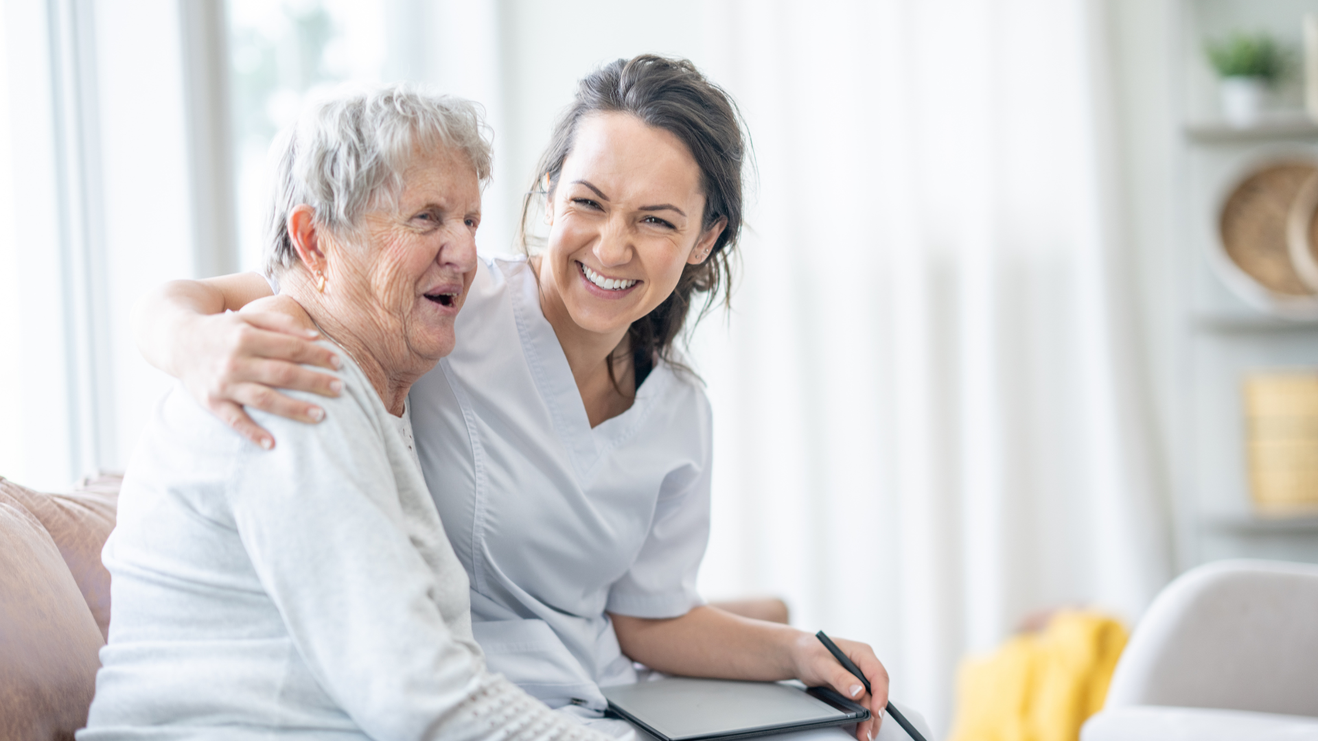 Smiling woman sitting with an elderly person on a couch in a bright living room