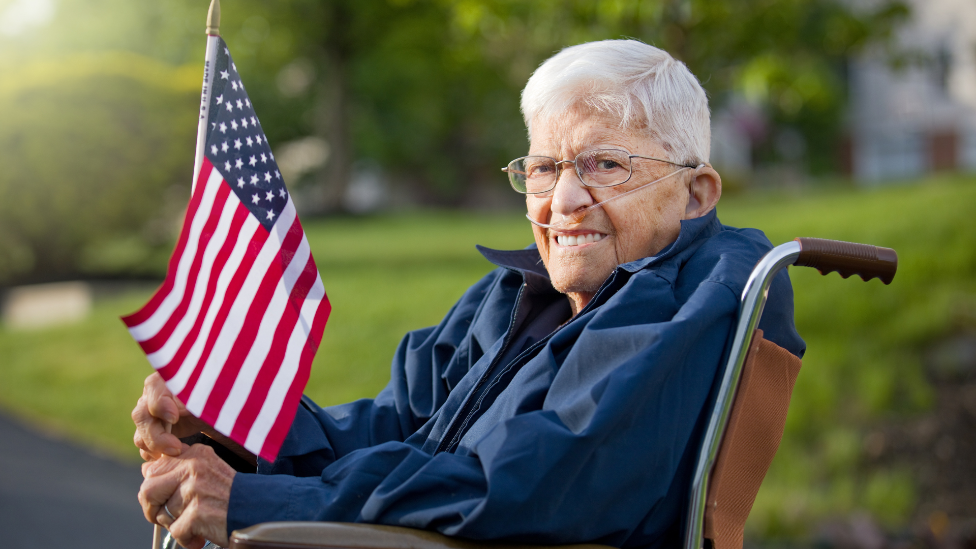 Smiling elderly man holding a small American flag outdoors on a lawn.