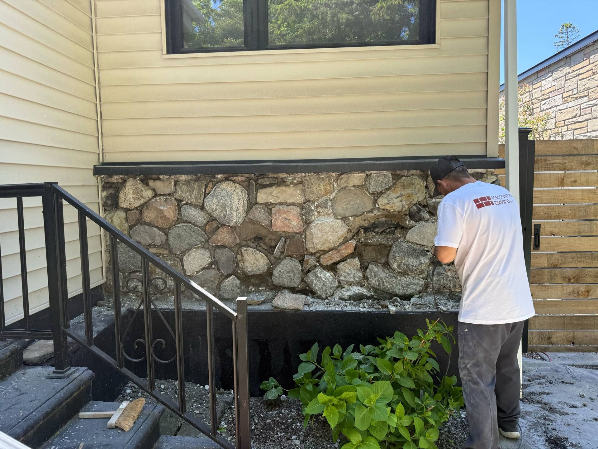 Une personne travaille à côté des fondations en pierre, sous le revêtement extérieur de la maison, près de la rampe d'escalier noire.