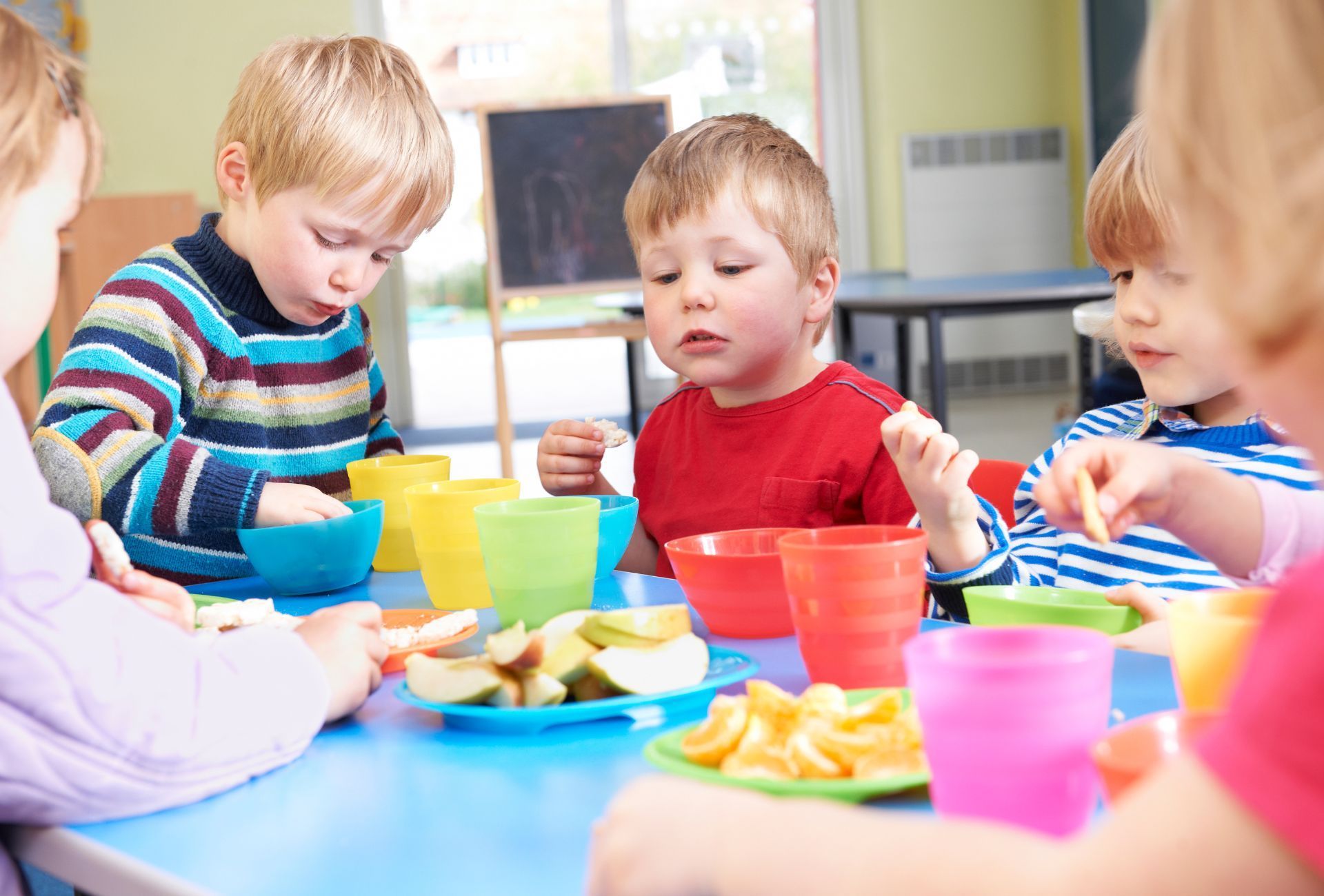Children eating fruit at a blue table in a classroom with colorful cups.