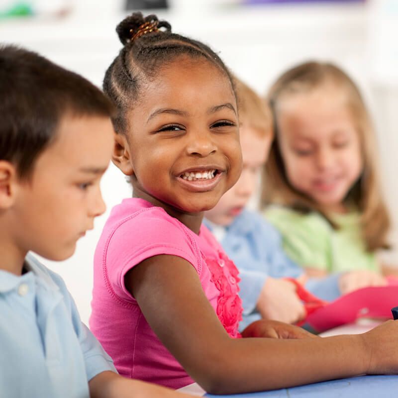 Smiling child in pink shirt, looking at the viewer, with other children at a table.