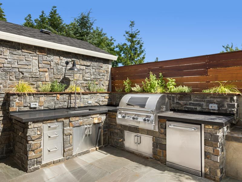 Stone outdoor kitchen with grill, sink, and mini fridge beside a stone wall and wooden fence