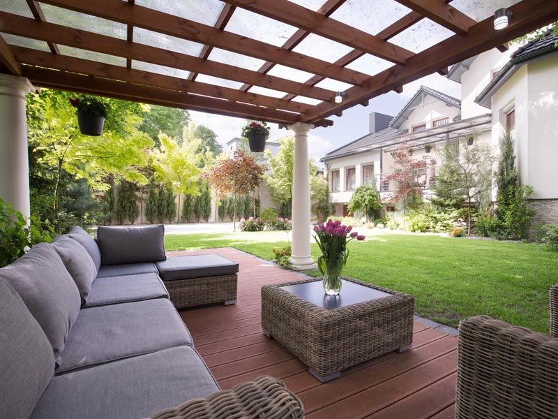 Covered patio with gray outdoor seating, wicker tables, and a green lawn beside a large house.