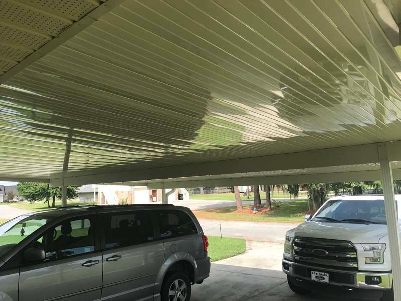 Two parked vehicles under a corrugated metal carport, with a grassy yard and houses in the background.